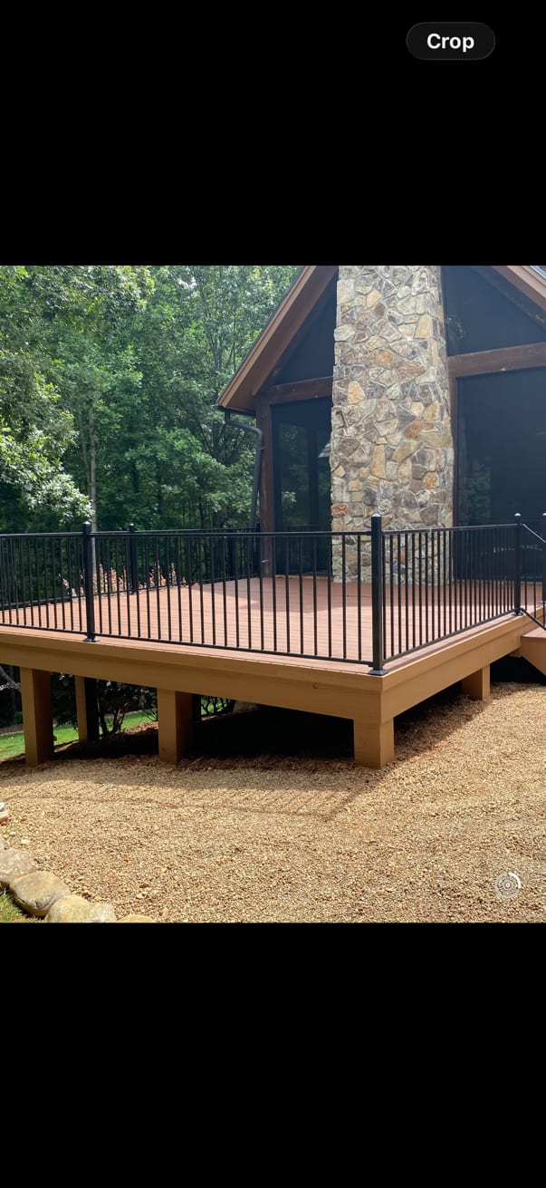Raised timber deck with black metal balusters built off the back of a house, set against a full-height stone chimney with trees behind.