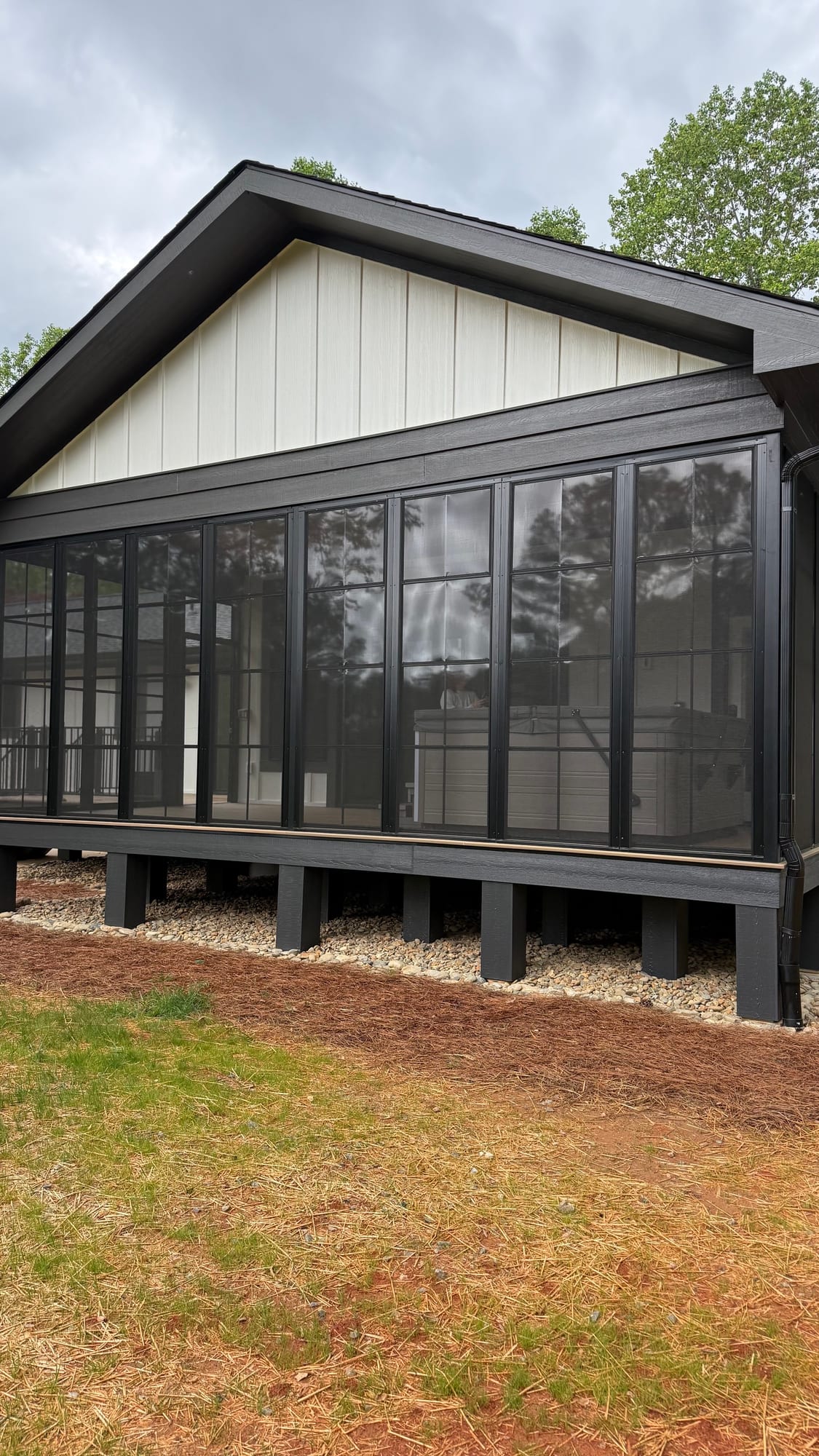 Screened porch running the length of a white board-and-batten house with black trim, raised on painted posts above a mulched bed.