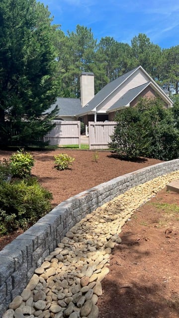Curved segmental stone retaining wall following the contour of a slope, with a river-rock swale at its foot and mulched beds above.