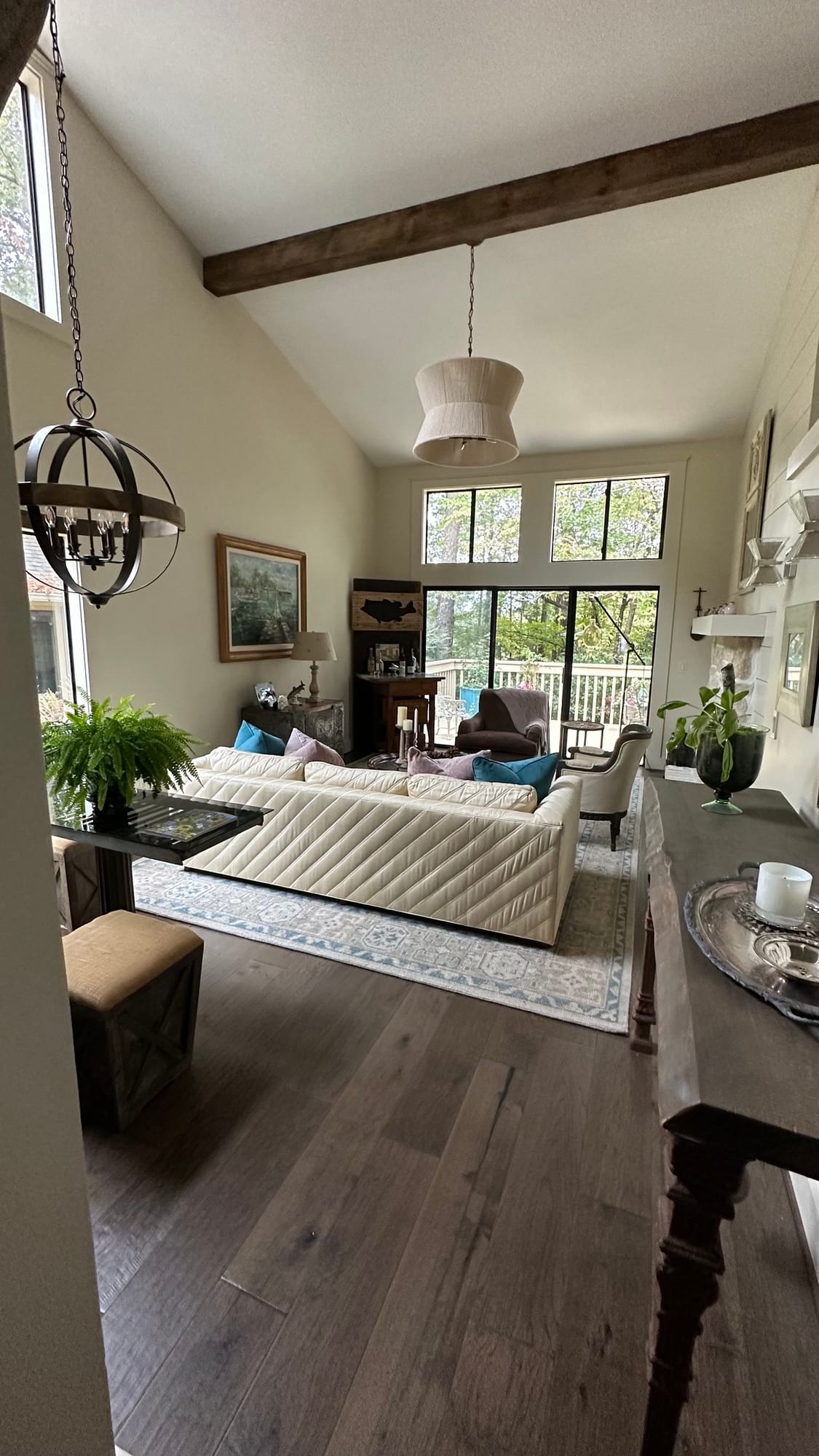 Vaulted living room with an exposed beam and orb chandelier, looking across seating to full-height windows and dark hardwood floors.