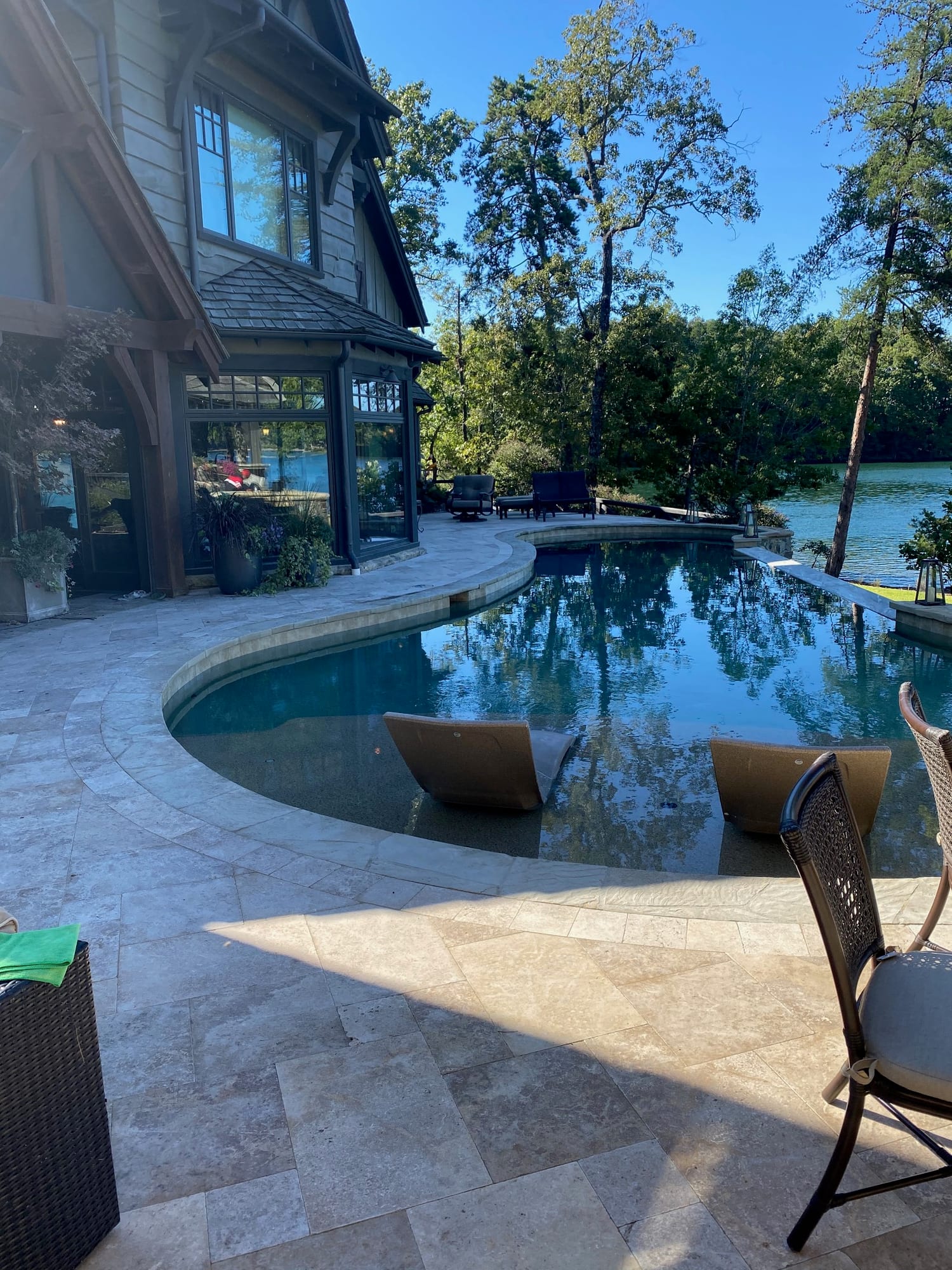 Freeform pool with travertine surround curving past a timber-frame lake house, with the lake beyond the far coping.